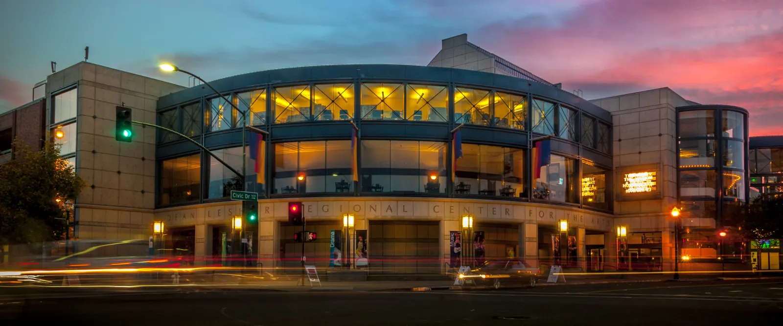 The Lesher Center for the Arts at dusk: a curved glass rotunda lit from within above a stone frieze, with car light trails on Civic Drive.